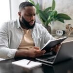 man-reviewing-documents-while-working-on-laptop