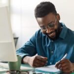 smiling-man-writing-notes-at-desk-with-computer