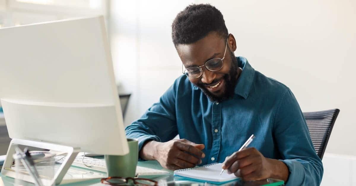 smiling-man-writing-notes-at-desk-with-computer
